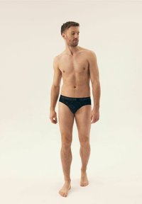 Czarne slipki z teksturowanym wzorem, z kontrastującym czarnym paskiem z napisem "HENDERSON". Model ma neutralną postawę i jest boso.