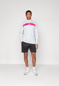 Lys grå sportsjakke med glidelås og rosa aksenter, sammen med sorte shorts. Har høy krage og elastisk midje. Hvite treningssko.
