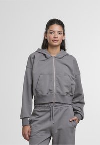 Haut à capuche zippé gris en tissu doux, avec une capuche, des poches avant et des poignets à côtes. Assorti à un pantalon de jogging à cordon de serrage.