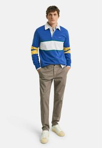 Camisa de rugby azul con rayas blancas, amarillas y verdes, combinada con pantalones beige y zapatillas crema. Ajuste casual con cuello.