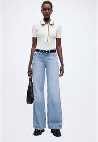 Εκρού μπλουζάκι με γιακά και λεπτομέρειες κουμπιών, σε συνδυασμό με ανοιχτό μπλε τζιν παντελόνι φαρδιάς γραμμής. Μαύρη ζώνη και χοντρά μαύρα παπούτσια. Μινιμαλιστική τσάντα.