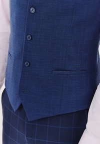 Granatowa lniana kamizelka w kwadratowym kroju, z sześcioma guzikami i jedną kieszenią z przodu. Założona na jasnoróżową koszulę i spodne w kratę.