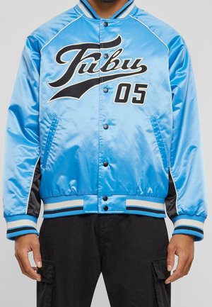 Mann trägt eine hellblaue Satin-Collegejacke mit schwarz-weiß gestreiften Bündchen und dem Schriftzug "Fubu 05" auf der Brust, kombiniert mit schwarzen Hosen.