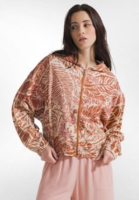 Frau trägt eine pinke und orange Blättermuster-Reißverschlussjacke mit elastischen Bündchen und hellrosa Hose, schaut zur Seite.