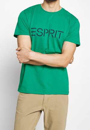 Man with arms crossed wearing a green Esprit T-shirt and beige pants against a plain white background.