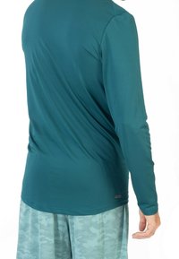 Camisa de manga larga color teal con una textura suave. Combinada con pantalones cortos de camuflaje en un tono azul claro. Color uniforme con un detalle de marca mínimo.