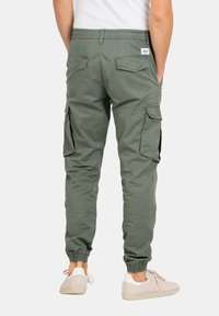Pantalones cargo verdes con cintura y puños elásticos, que cuentan con bolsillos laterales y detalles en el bolsillo trasero. Llevados con zapatillas de deporte de color claro.