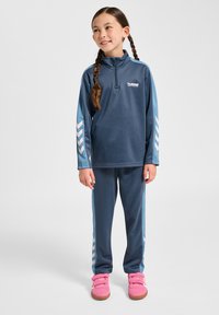 Blauwe trainingspak gemaakt van zacht materiaal, met een ritskraag en chevronaccenten. Combinatie met roze klittenbandschoenen.