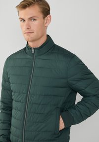 Zelená vyp padded bunda s vysokým golierom, s horizontálne prešívanou textúrou, plným zipsom a bočnými vreckami. Hladká povrchová úprava textilu.