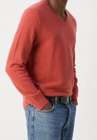 Un homme portant un pull en V couleur rouille, les mains dans les poches, associé à un jean bleu et une ceinture noire.