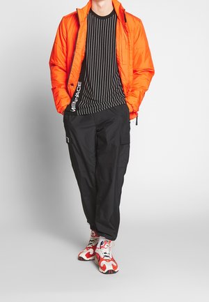 Orangen, gesteppte Jacke über einem schwarz gestreiften Hemd. Schwarze Cargo-Hosen und rot-weiße Sneakers mit schwarzen Sohlen.