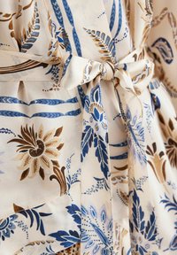Cremefarbener Stoff mit komplexen blauen und braunen Blumenmustern, versehen mit einer gebundenen Schleifendetails. Glatte Textur mit Falten, die das Design betonen.