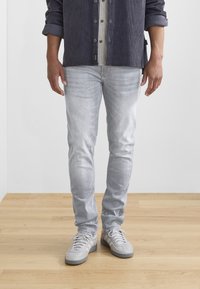 Světlé šedé slim-fit džíny z denimové látky, s jemným vyblednutím a detaily šití, v kombinaci se světlými šedými teniskami na dřevěné podlaze.