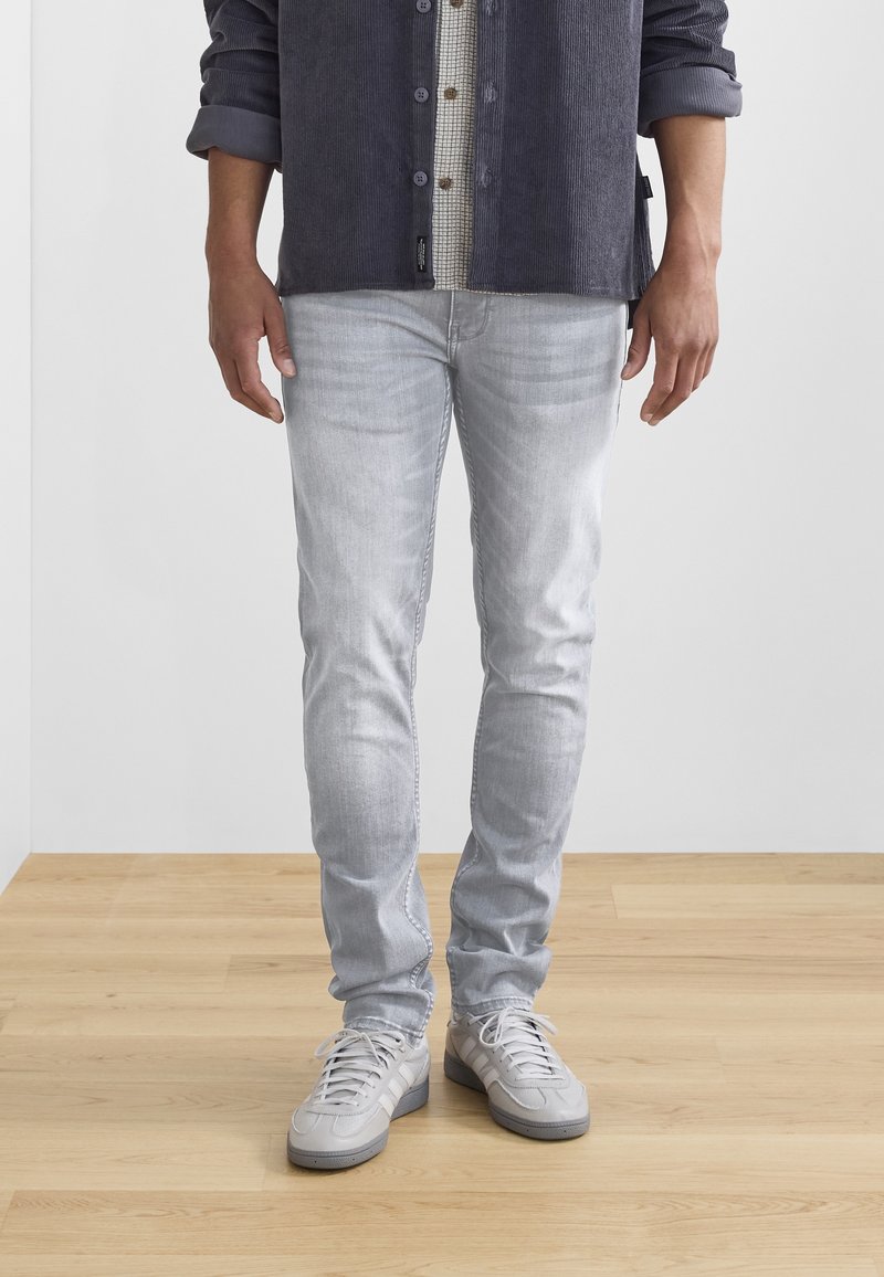 Světlé šedé slim-fit džíny z denimové látky, s jemným vyblednutím a detaily šití, v kombinaci se světlými šedými teniskami na dřevěné podlaze.