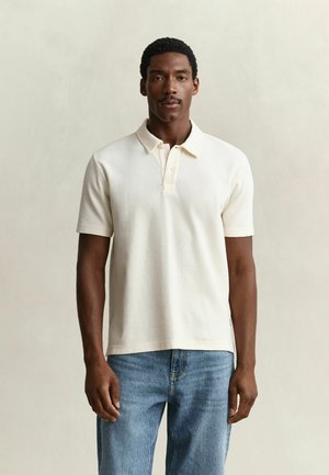 Homme portant un polo blanc à manches courtes et un jean bleu, debout devant un fond beige uni.
