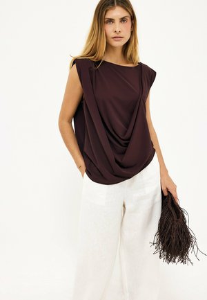 Woman wearing a sleeveless dark draped top and loose white pants, holding a dark fringed handbag in one hand against a plain background.