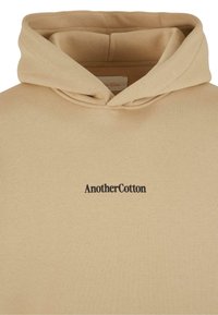 Beige katoenen hoodie met een voorste kangoeroezak en een zwart "AnotherCotton" logo op de borst. Zachte textuur en aanpasbare trekkoorden in de capuchon.