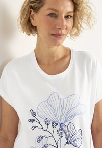 Hvid t-shirt med korte ærmer, prydet med et blåt blomsterlinjeteckning. Rund halsudskæring og en afslappet pasform. Glat bomuldsstruktur.