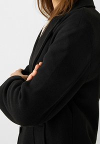 Svart ullkappa med texturerad yta och skräddarsydd passform. Har en kavajkrage med notch och dold knappstängning. Synliga sömmar förstärker strukturen.