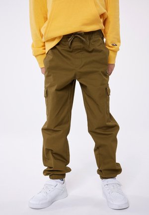 Persona che indossa pantaloni cargo verde oliva con polsini elasticizzati, una felpa gialla e sneakers bianche, in piedi contro uno sfondo semplice.