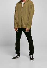 Oliwkowa zielona prążkowana bluza z kapturem zapinana na zamek, połączona z czarnymi dżinsami i czarnymi trampkami z białymi akcentami. Widoczny detal tekstury tkaniny.