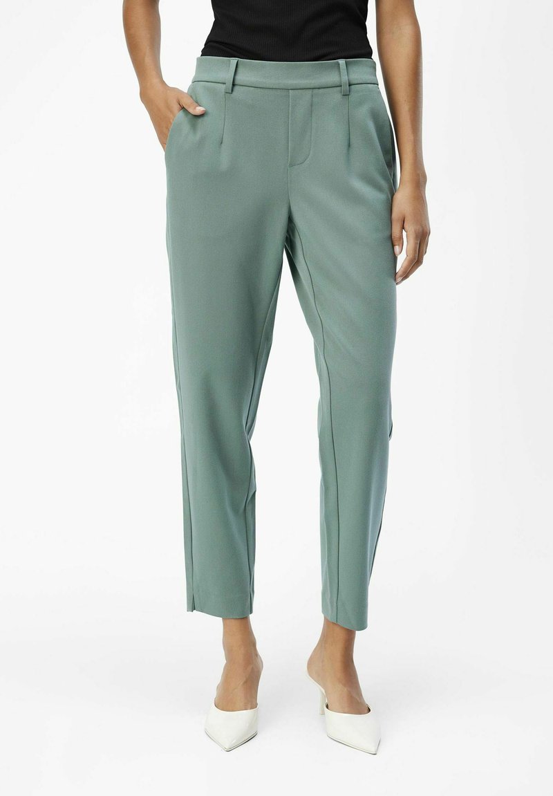 Pantalons ajustés vert clair avec une taille plate, des poches sur les côtés et une coupe fuselée, le tissu semble lisse et légèrement brillant.