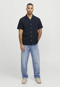 Marineblauw overhemd met korte mouwen, een zak, knoopsluiting en een ontspannen pasvorm, gecombineerd met lichtblauwe wijde jeans en beige schoenen.