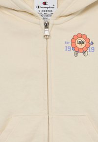 Felpa con zip di colore beige chiaro realizzata in morbido tessuto. Presenta una grafica di un fiore stilizzato e la scritta "EST. 1919". Include tasche frontali e cappuccio.