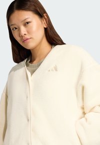 Krämfärgad fleecejacka med avslappnad passform, featuring en v-ringning, knäppning fram och en subtil logo på vänster bröstområde.