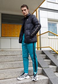 Sort pufjakke med logo, turkise træningsbukser med sorte striber, og sorte og hvide sneakers. Model poserer på betontrapper.