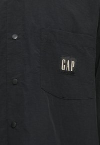 Svart skjorte med knapper og brystlomme, med en sydd svart etikett med hvit "GAP" tekst.