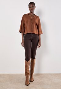 Brun cropped button-up skjorte med V-udskæring, parret med mørkebrune tætsiddende jeans og beige knæ-høje støvler med spidse tæer.