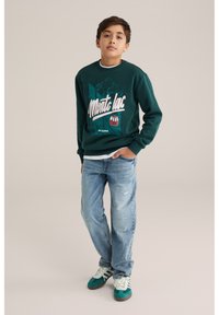 Zaļš sporta krekls ar kalna grafiku un baltu tekstu, apvienots ar valkātiem ziliem džinsiem un zaļām sporta kedām ar baltām šņorēm.