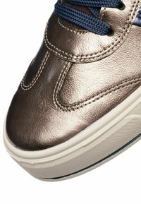 Detailní záběr na kovově bronzovou špičku tenisky s modrými tkaničkami a bílou texturovanou gumovou podrážkou.