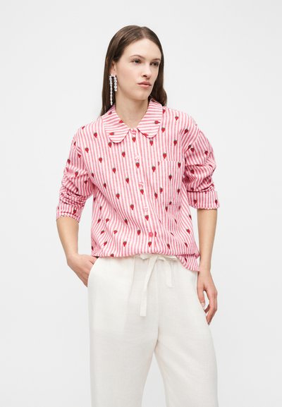 Woman wearing long-sleeve pink and white striped button-up shirt with small red strawberry prints and white drawstring pants, standing with hands in pockets.