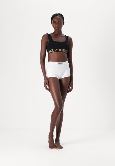 Schwarzes Crop-Top mit goldenen geometrischen Mustern und einem runden Logo, kombiniert mit weißen, hochgeschnittenen Unterhosen mit einem Etikett. Beide Kleidungsstücke sind glatt.