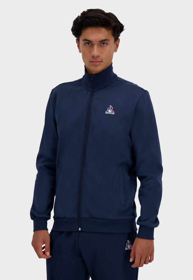 Chaqueta de chándal azul marino con cremallera, fabricada en un tejido suave, con cuello alto, puños acanalados, dos bolsillos laterales y detalle de logo en el lado izquierdo del pecho.