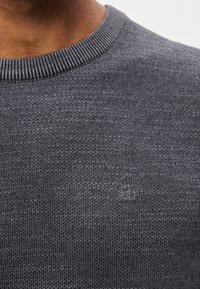 Ciemnoszary sweter dzianinowy z prążkowanym dekoltem, z subtelnym wytłoczonym logo z przodu. Tekstura jest gładka i równa.