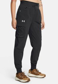 Pantalones deportivos negros con cintura el�ástica, bolsillo lateral con cremallera y bolsillo tipo cargo. Hechos de material ligero. Combinados con zapatillas marrones.
