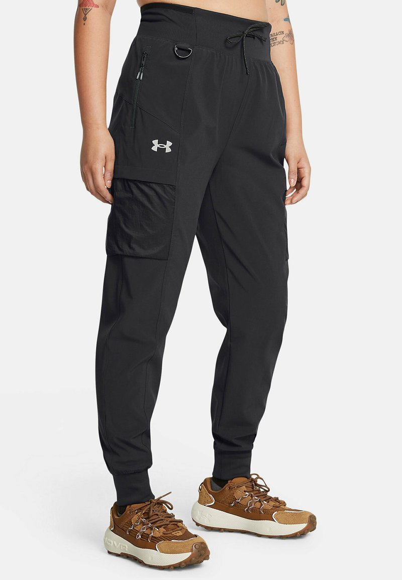 Pantalones deportivos negros con cintura elástica, bolsillo lateral con cremallera y bolsillo tipo cargo. Hechos de material ligero. Combinados con zapatillas marrones.