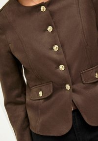 Veste marron en matériau doux avec un col rond, quatre boutons décoratifs en or et deux poches avant avec des accents de boutons.