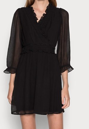 Femme portant une robe noire mi-genou avec des manches longues en voile transparent, un décolleté en V à volants et une ceinture texturée.