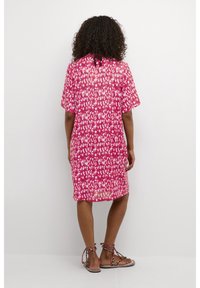 Kvinna med lockigt hår som bär en knäkort klänning med rosa och vita mönster samt sandaler med platt sula, stående vänd mot en vit vägg.