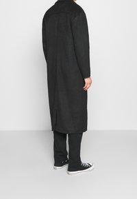 Persona che indossa un lungo cappotto nero, pantaloni neri e sneakers bianche e nere, in piedi contro una parete e un pavimento grigi semplici.