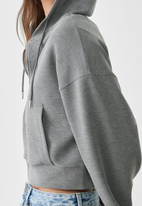 Grijze korte hoodie met een ritssluiting aan de voorkant, voorzien van een capuchon, grote zakken en lange mouwen. Gemaakt van zachte, gestructureerde stof met een casual ontwerp.