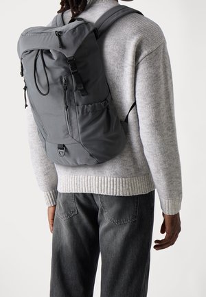 Sac à dos gris au design arrondi, texture en tissu, bretelles ajustables et fermeture à cordon de serrage. Porté avec un pull gris et un jean.