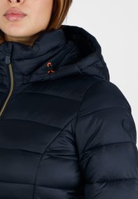 Giacca imbottita navy con colletto alto e cappuccio. Caratterizzata da sezioni trapuntate, una zip dorata e un dettaglio in arancione sullo stringi vita. Tessuto testurizzato.
