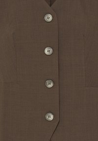 Tissu marron avec quatre gros boutons ronds à quatre trous disposés verticalement sur le devant d’un vêtement sur mesure.