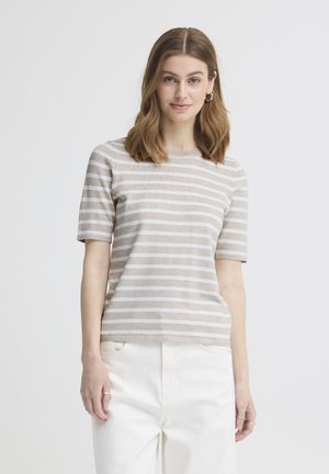 Žena se světlými hnědými vlasy, oblečena v béžovém a bílém pruhovaném tričku s krátkými rukávy a bílých kalhotách, stojící před jednobarevným pozadím.