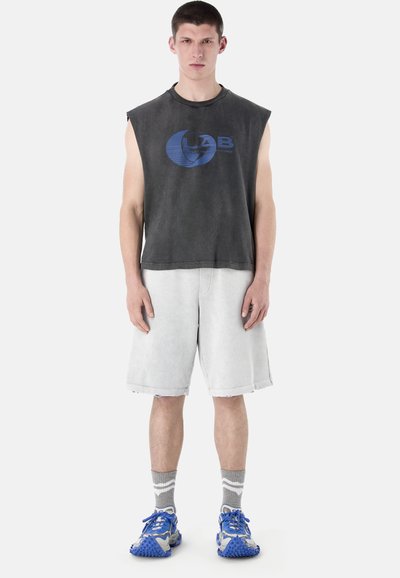 Jeune homme portant un débardeur noir à motif, un short gris clair arrivant aux genoux, des chaussettes grises et des chaussures de sport bleues, debout face à l'avant.